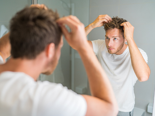 Hair loss man looking in bathroom mirror putting wax touching his hair styling or checking for hair loss problem. Male problem of losing hairs.